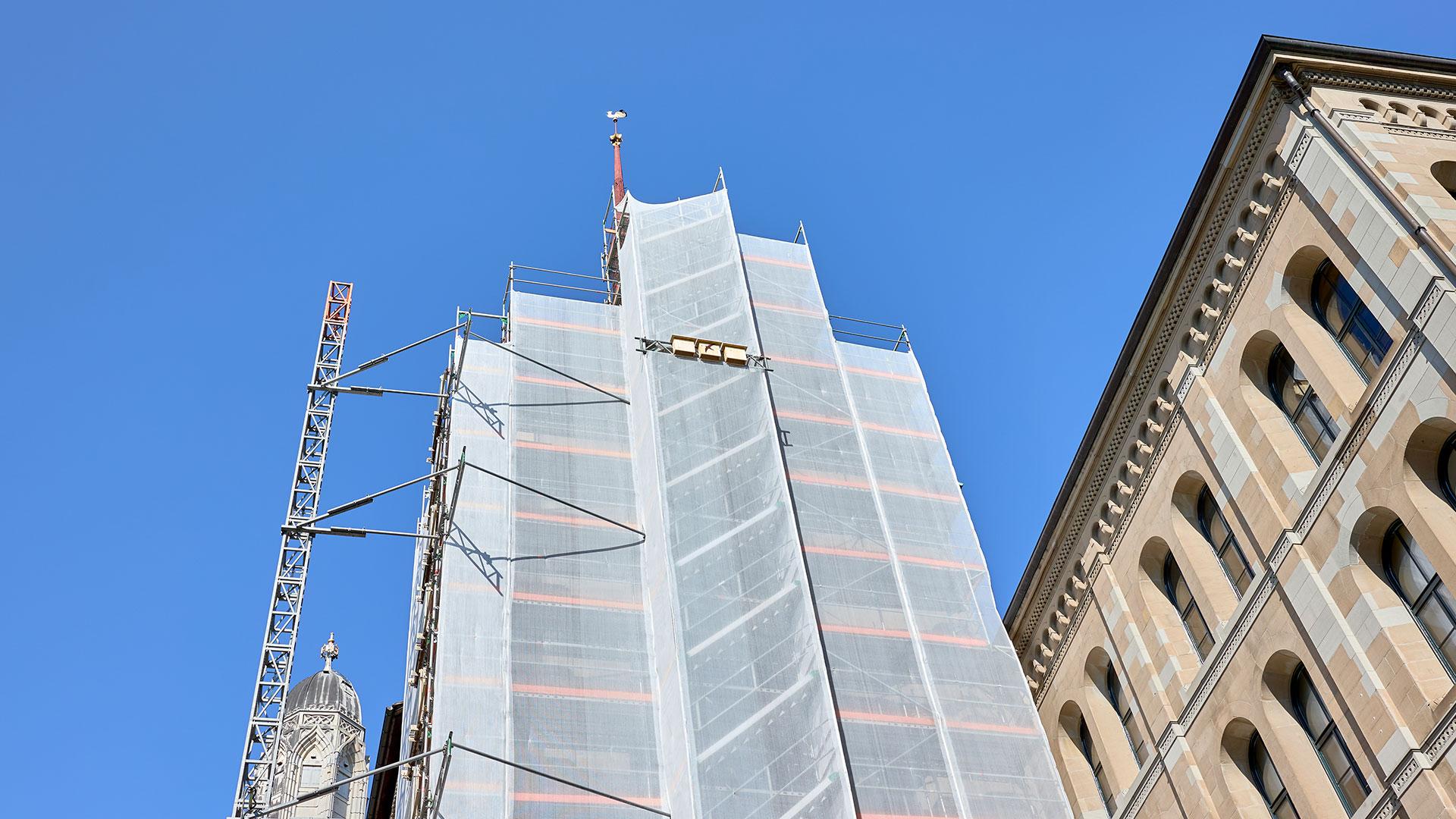 Ein hoher Kirchturm, teilweise mit Baugerüst und weißer Plane verdeckt, neben einem verzierten Gebäude unter blauem Himmel.