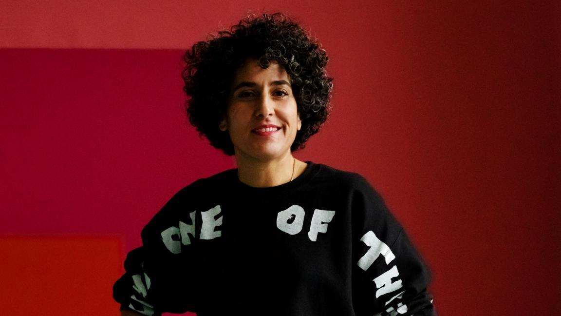 Eine Frau mit lockigem, dunklem Haar und einem schwarzen Pullover mit weisser Aufschrift posiert vor einem zweifarbigen Hintergrund. Sie hat die Hände in die Hüften gestützt und lächelt leicht in die Kamera. Sie trägt goldene Armreifen am rechten Handgelenk und einen goldenen Ring. Der Hintergrund ist vertikal zweigeteilt, wobei die linke Seite in einem tiefen Pink und die rechte Seite in einem etwas helleren Rotton gehalten ist.