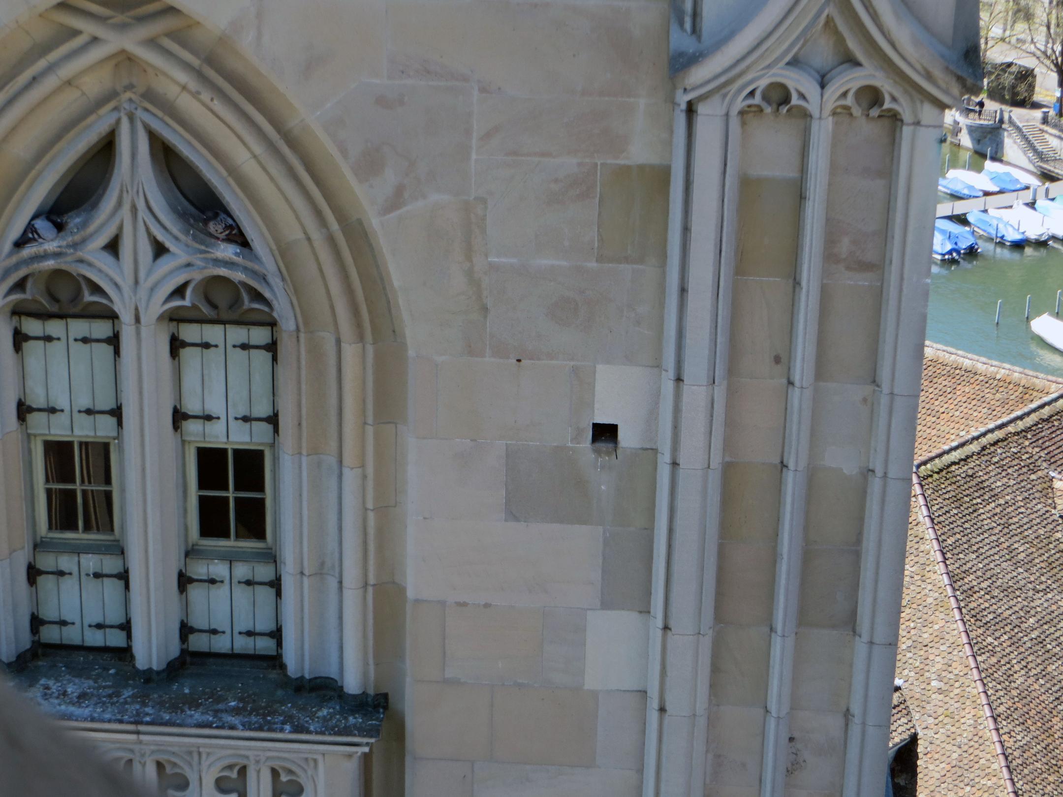 Eine Aussenaufnahme eines Gebäudes aus hellem Stein mit gotischer Architektur, einschliesslich eines Spitzbogens mit einem verzierten Fenster und einem kleinen Loch im Mauerwerk.