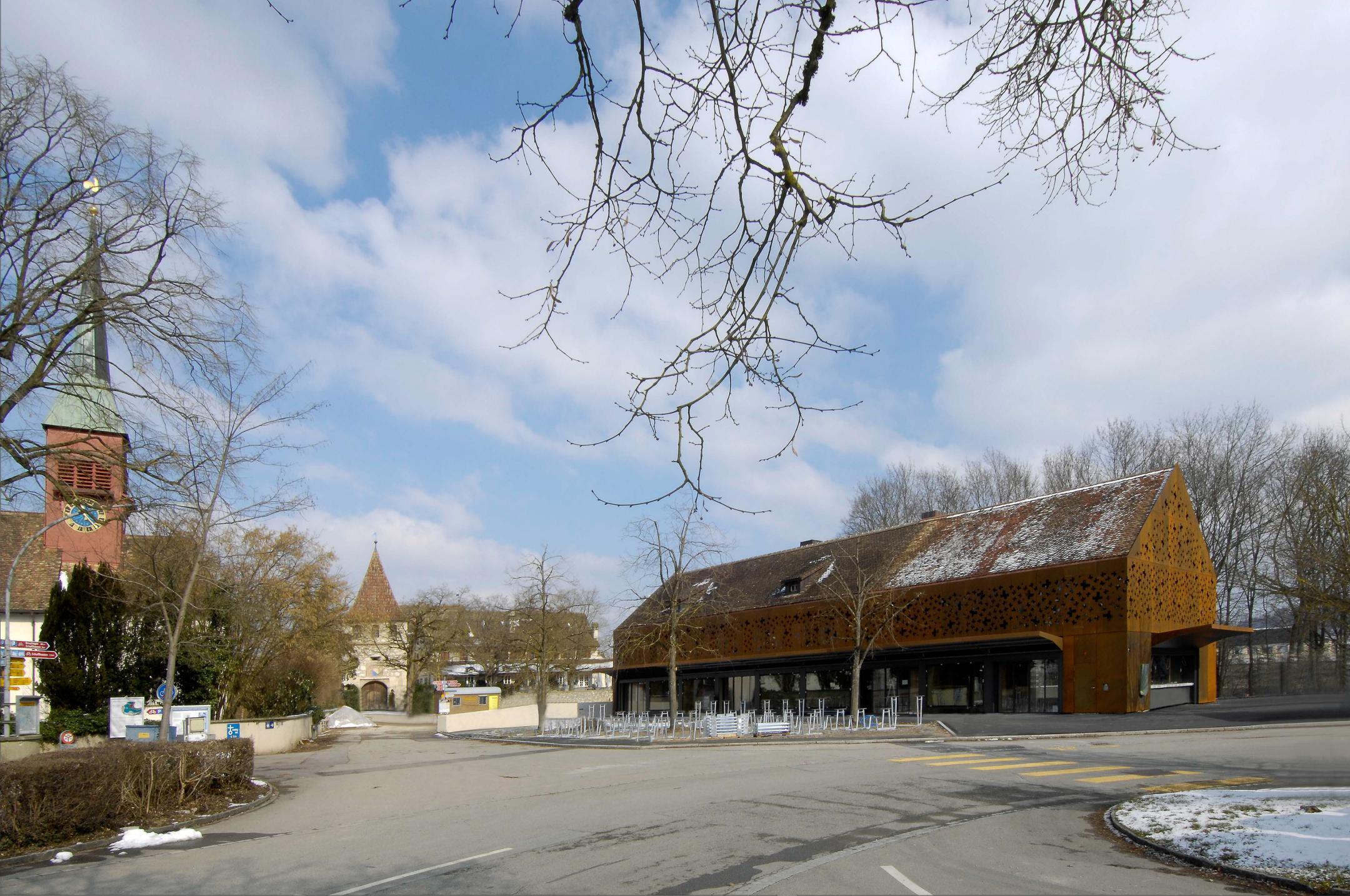 Ansicht an mehrere Gebäude über eine Strassenkreuzung fotografiert. Links eine ältere Kirche, in der Mitte ein Gebäude mit Turm und rechts ein neues Gebäude mit rostig wirkender Metallfassade.