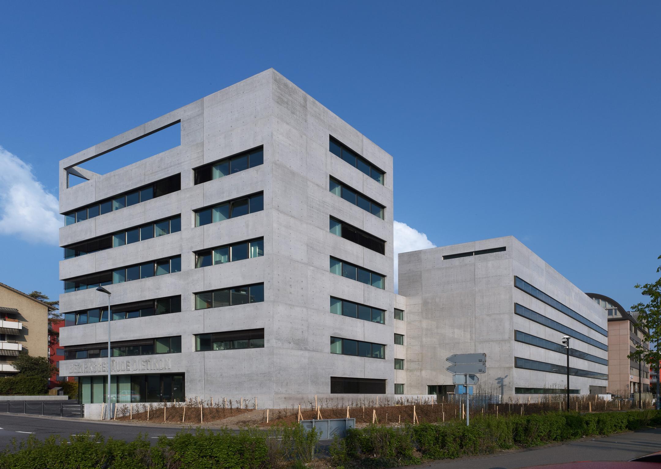 Bezirksgebäude in Sichtbeton vor blauem Himmel. Im Vordergrund die Strasse, gesäumt mit frisch gepflanzter Hecke.