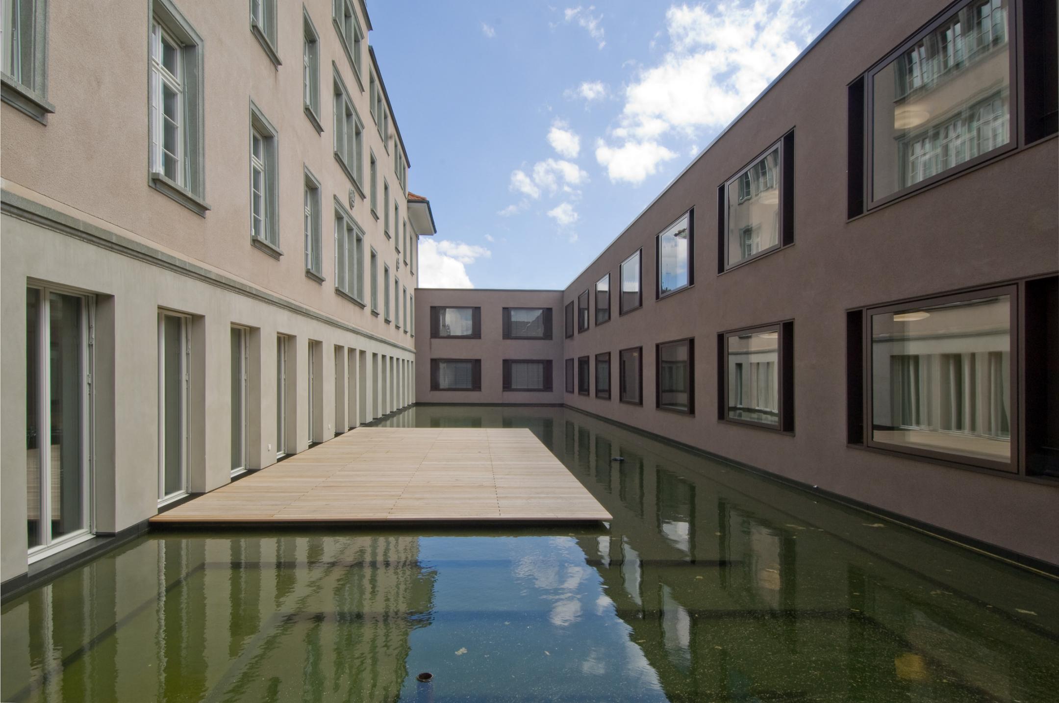 Links ein älterer Gebäudetrakt, rechts der angeschlossene neue Gebäudetrakt. Im entstandenen Hof ein kleiner Weiher mit begehbarem Holzfloss.