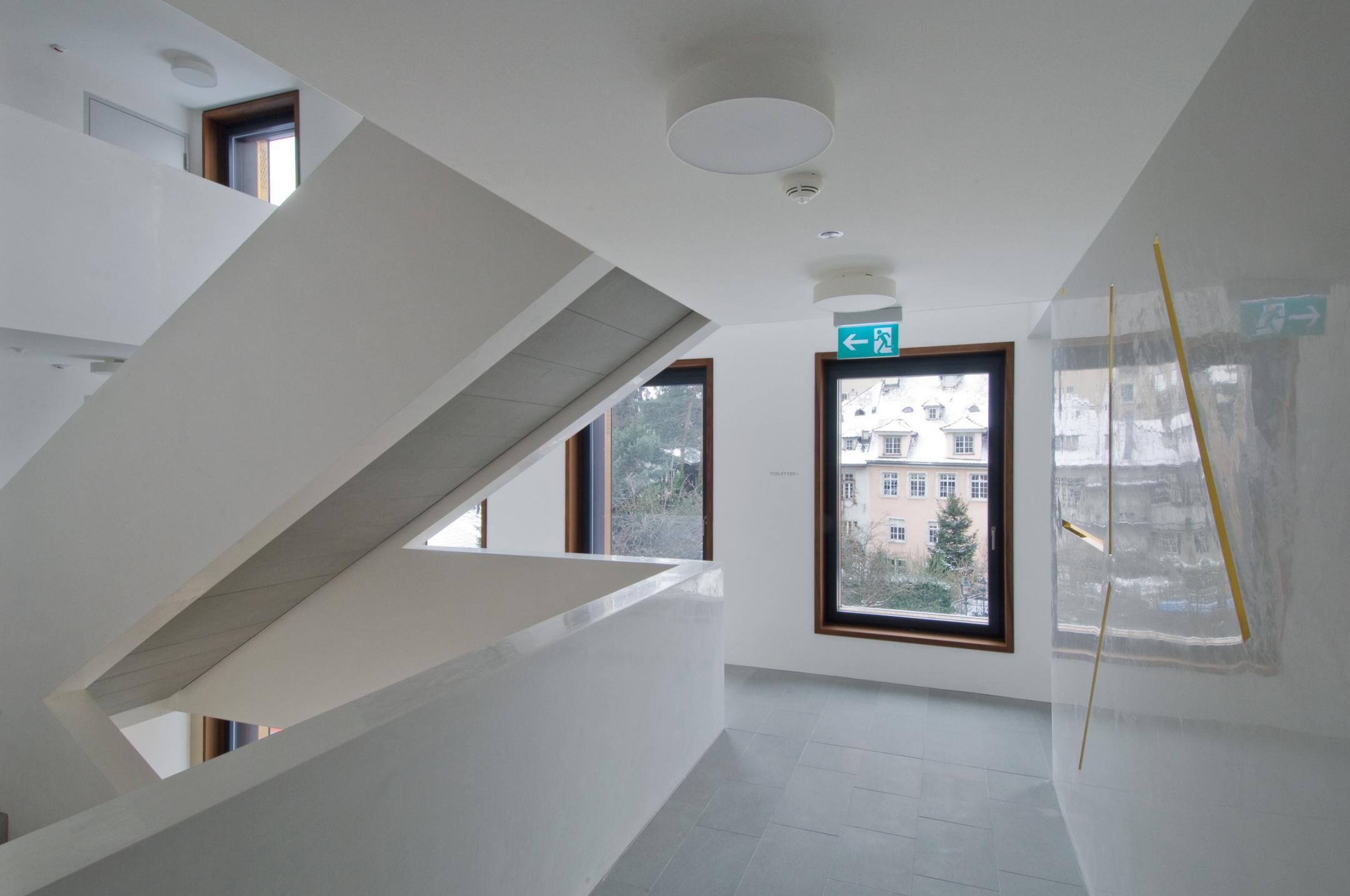 Innenansicht des weissen Treppenhauses. An der Wand sind vergoldete, keilschriftähnliche Metallstäbe montiert.