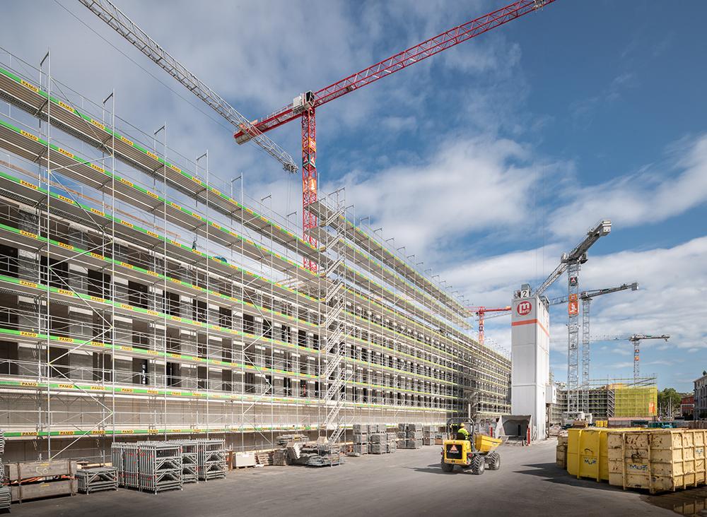 Ein in ein Baugerüst verpacktes Gebäude auf der PJZ-Baustelle im August 2018. Vier Kräne im Hintergrund, gelbe Baucontainer und eine Baumaschine. 