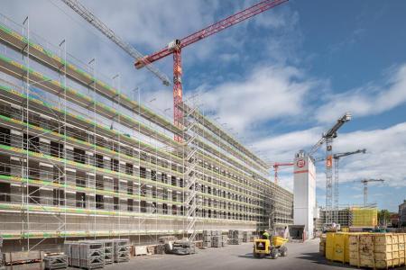 Ein in ein Baugerüst verpacktes Gebäude auf der PJZ-Baustelle im August 2018. Vier Kräne im Hintergrund, gelbe Baucontainer und eine Baumaschine. 