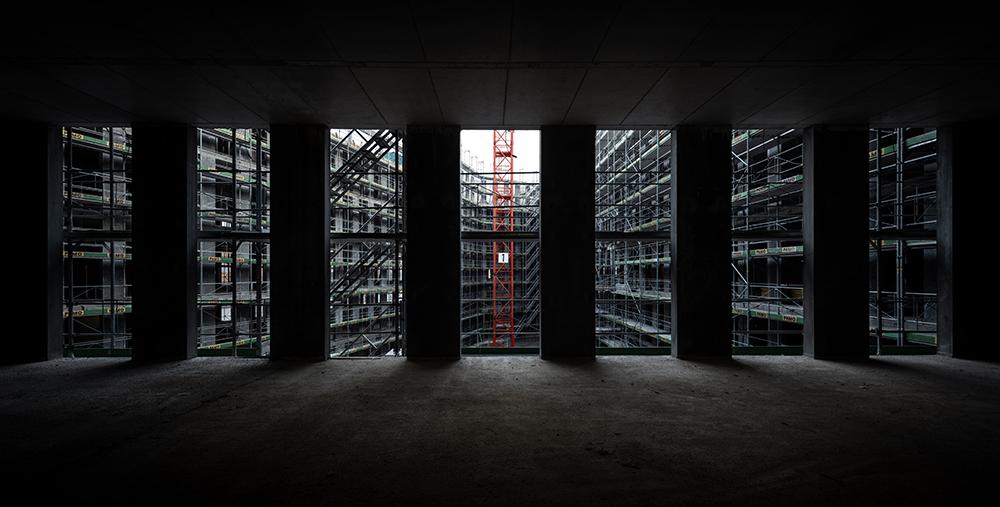 Sicht aus dem Innern der PJZ-Baustelle: Durch die Säulen des Gebäudes sieht man auf einen  von Baugerüsten eingerahmten Innenhof und den unteren Teil eines roten Krans. 