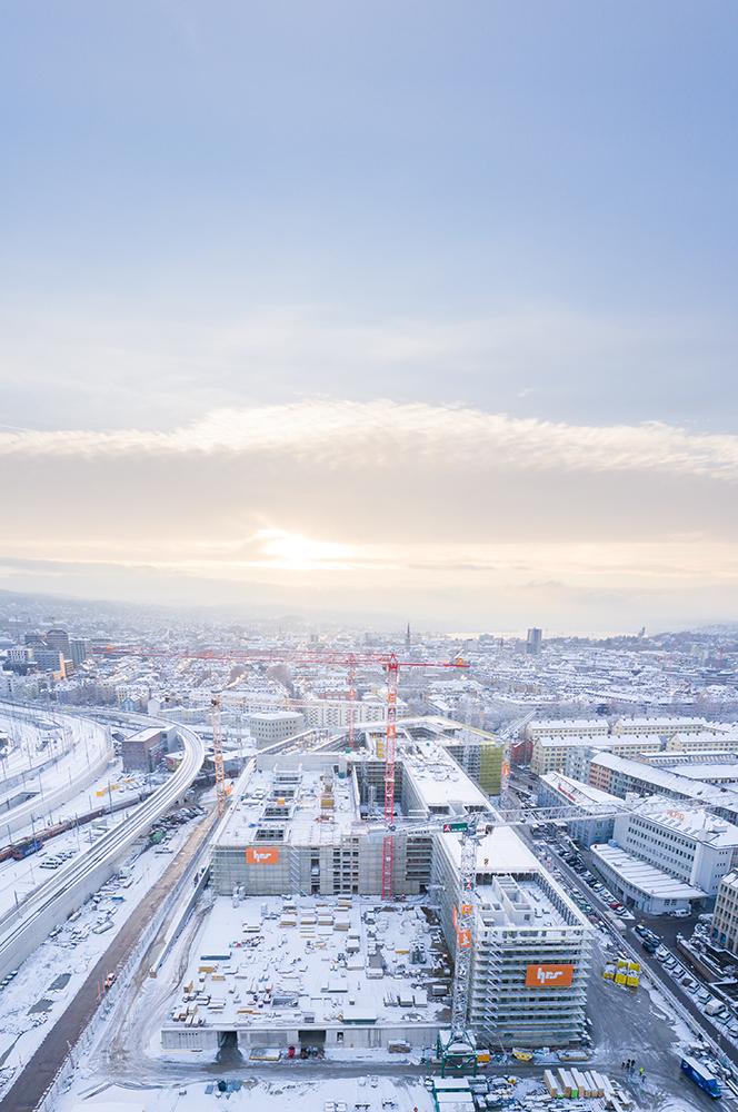 Die verschneite PJZ-Baustelle aus der Höhe aufgenommen, im Hintergrund ist das winterliche Panorama von Zürich sichtbar, bis hin zum Zürichsee. 