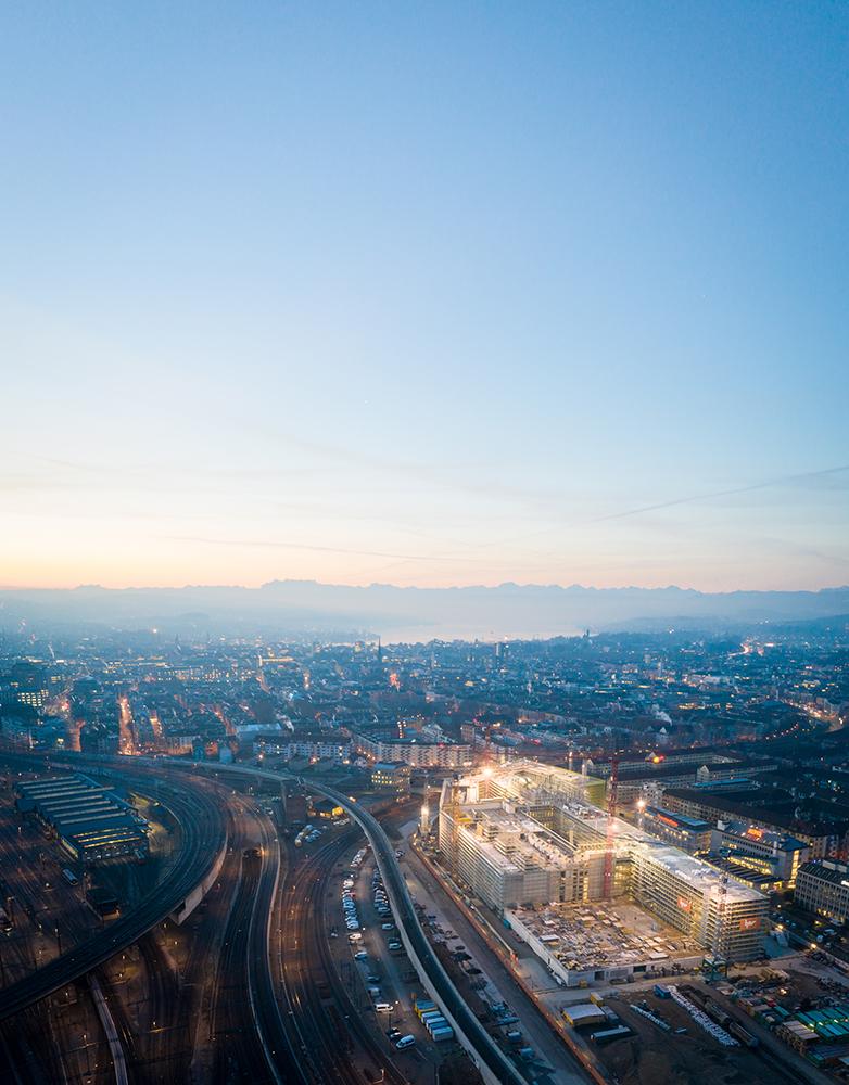 Die PJZ-Baustelle im Februar 2019: Die Baustelle liegt in der Dämmerung und ist beleuchtet, im Hintergrund die Stadt Zürich, der See und das Bergpanorama.