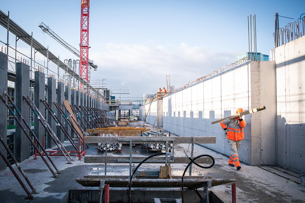 Die PJZ-Baustelle im Januar 2019: Ein Bauarbeiter in oranger Arbeitskleidung trägt ein Rohrelement über die Baustelle. 