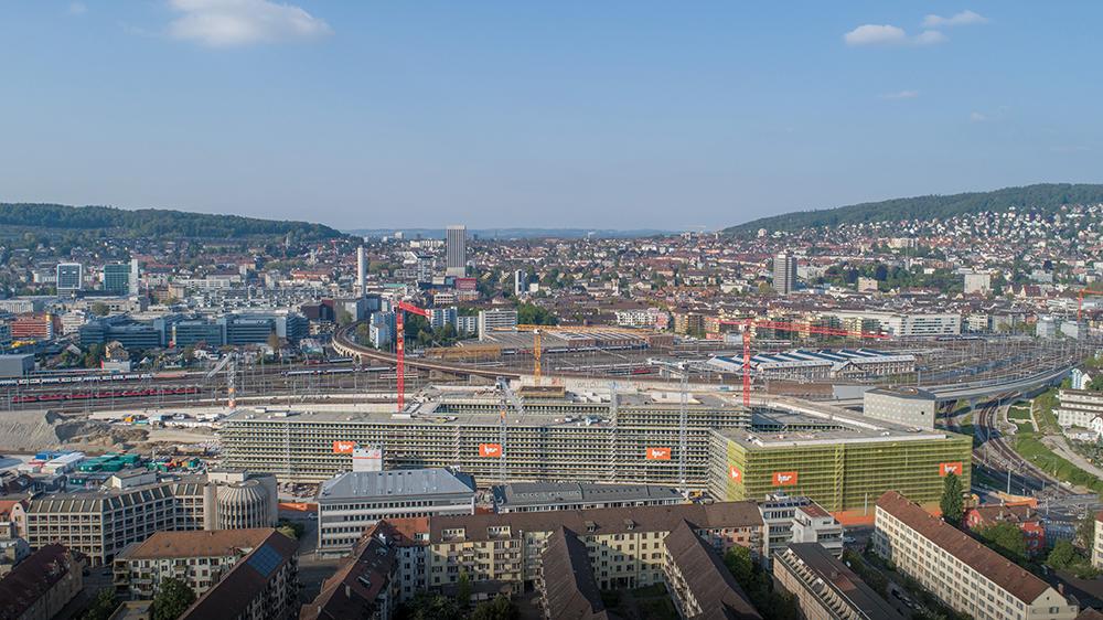 Die PJZ-Baustelle im Mai 2019, aus der Ferne und von schräg oben fotografiert. Im Vordergrund die Häuser der Hohlstrasse, dahinter Bahnschienen und die Stadt.