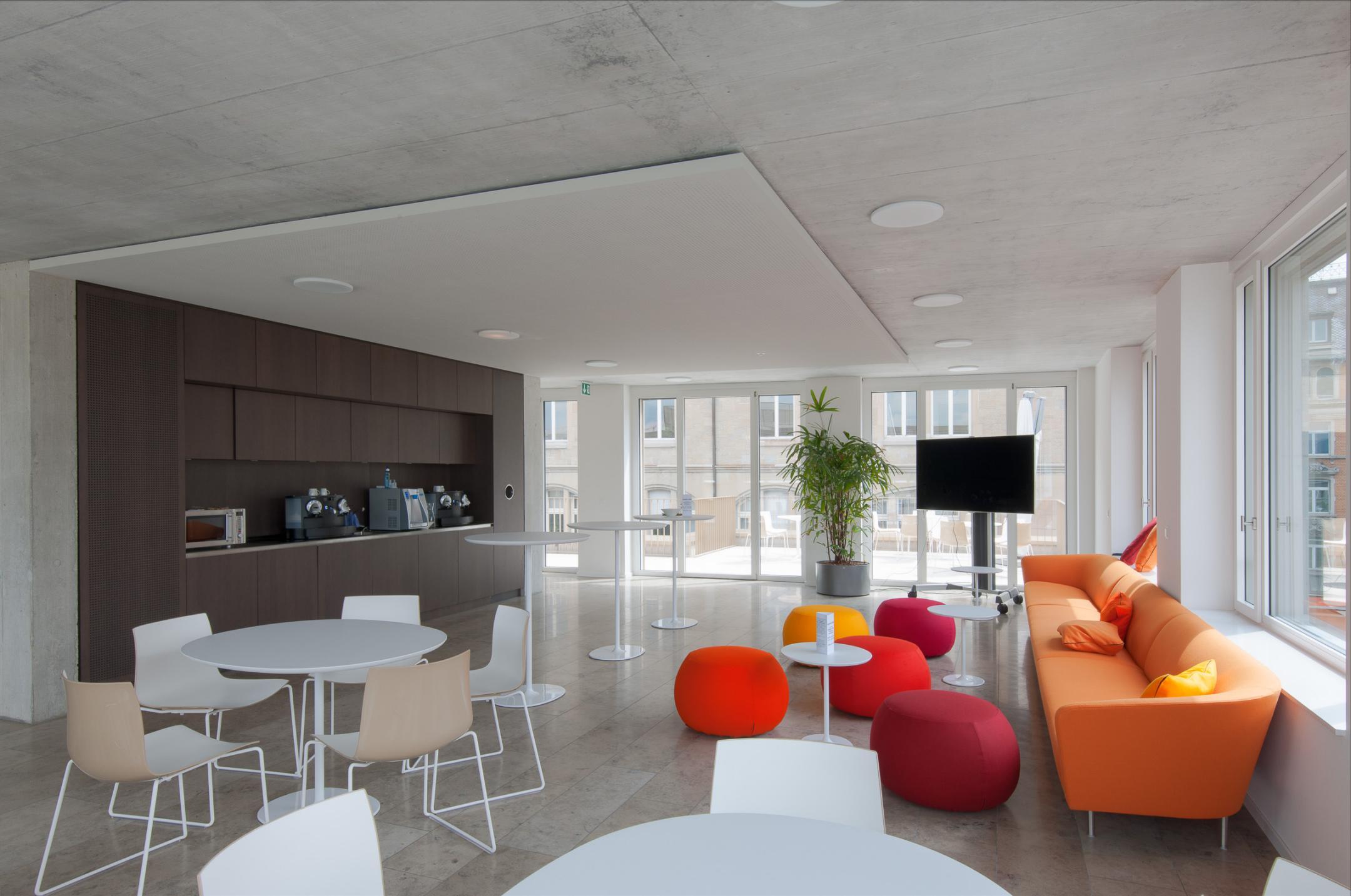 Sitzgelegenheiten aus kleinen Tischen mit Stühlen und einem Sofa ergänzt durch Sitzsäcke vor einer kleinen Küchenzeile.