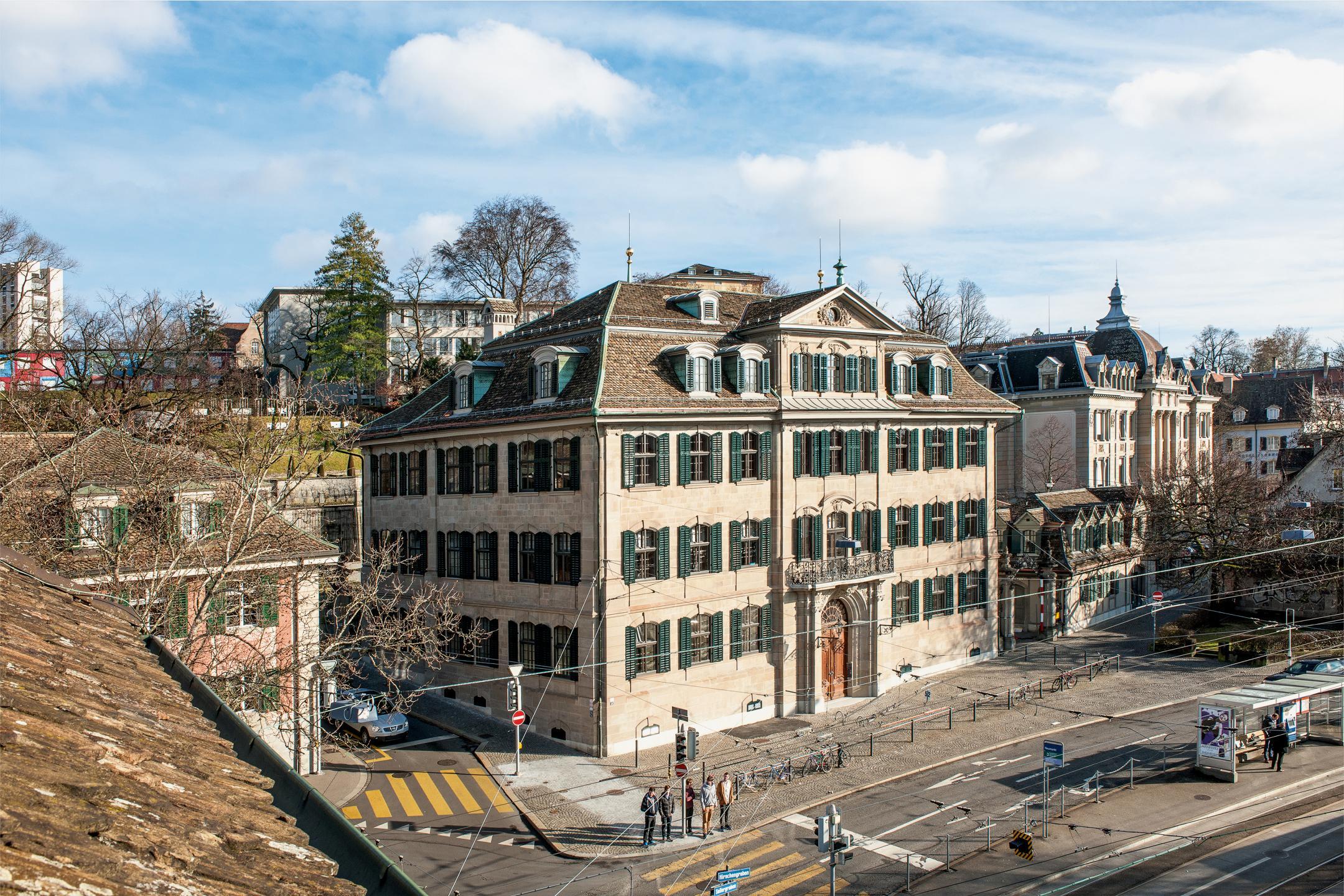 Strasse mit dem Rechberghaus von oben fotografiert. Personen stehen am Fussgängerstreifen. Der Himmel ist leicht mit Wolken durchzogen.