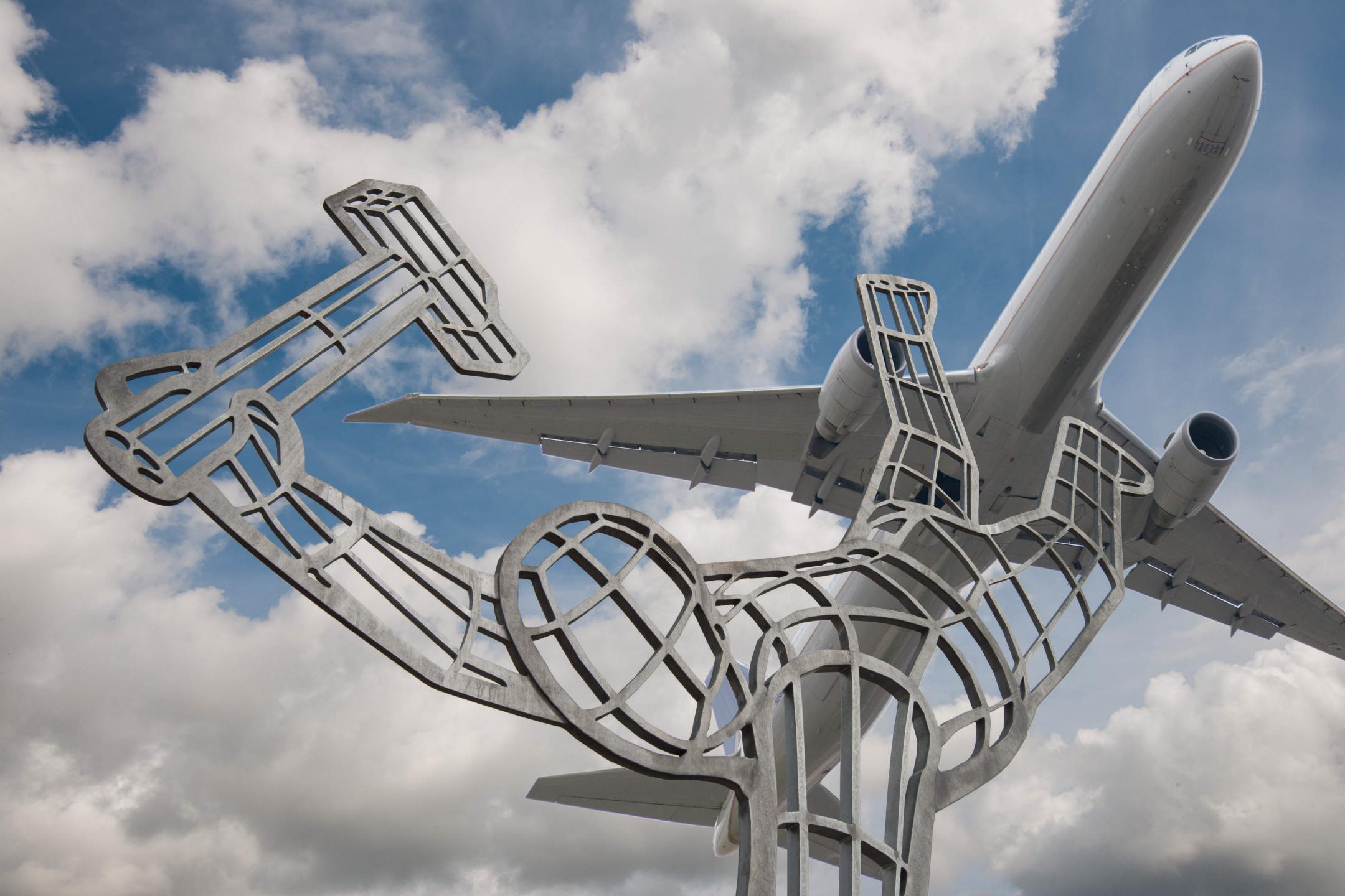 Stahlkonstruktion in Form eines Menschen, welcher auf einer Hand steht und in der zweiten Hand einen Hammer hält. Über der Stahlkonstruktion fliegt ein Flugzeug vor bewölktem Himmel.