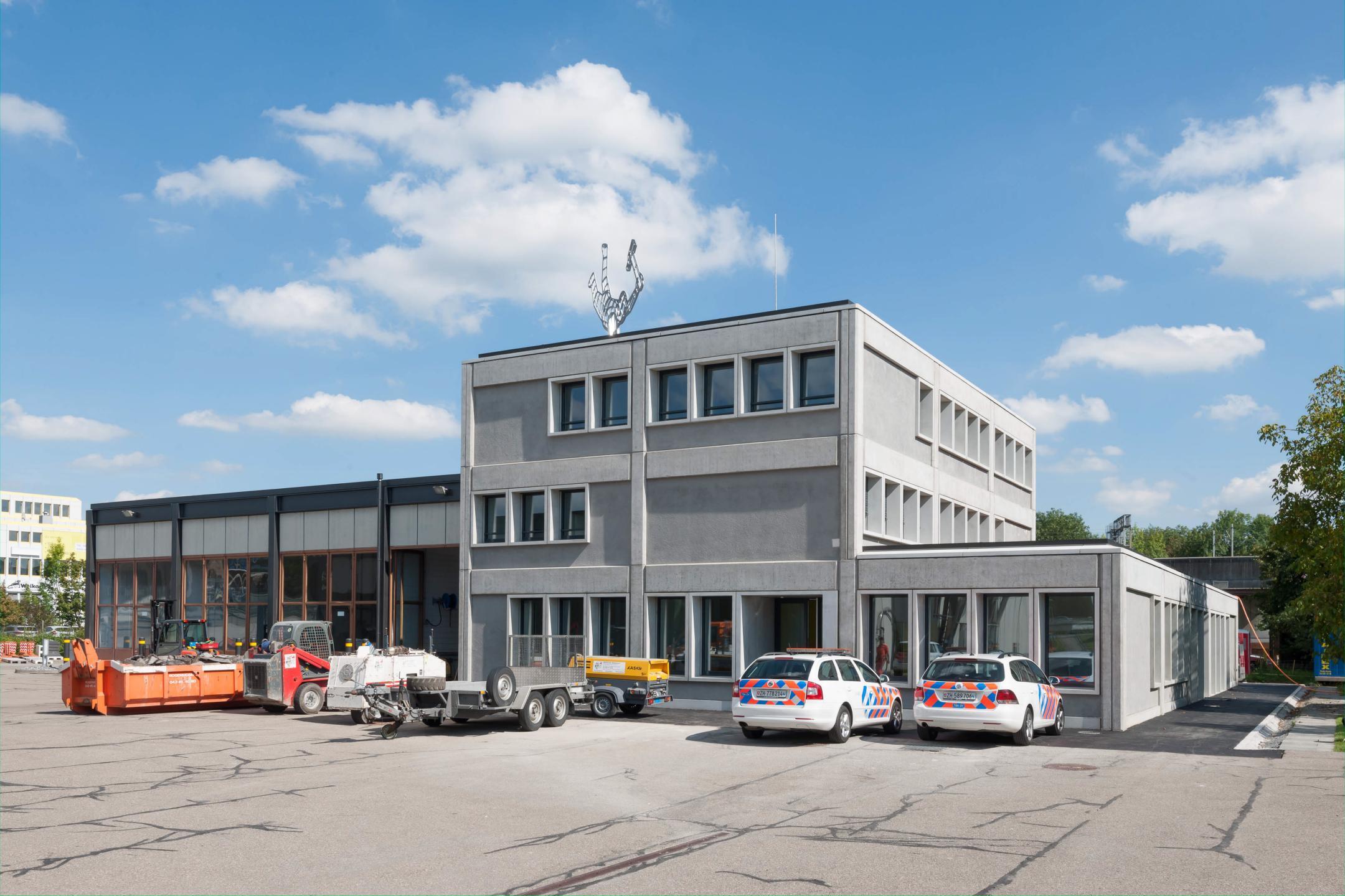 Gesamansicht eines Betongebäudes. Vor dem Gebäude sind mehrere Autos in auffälligen Tiefbauamt-Farben und Anhänger geparkt.