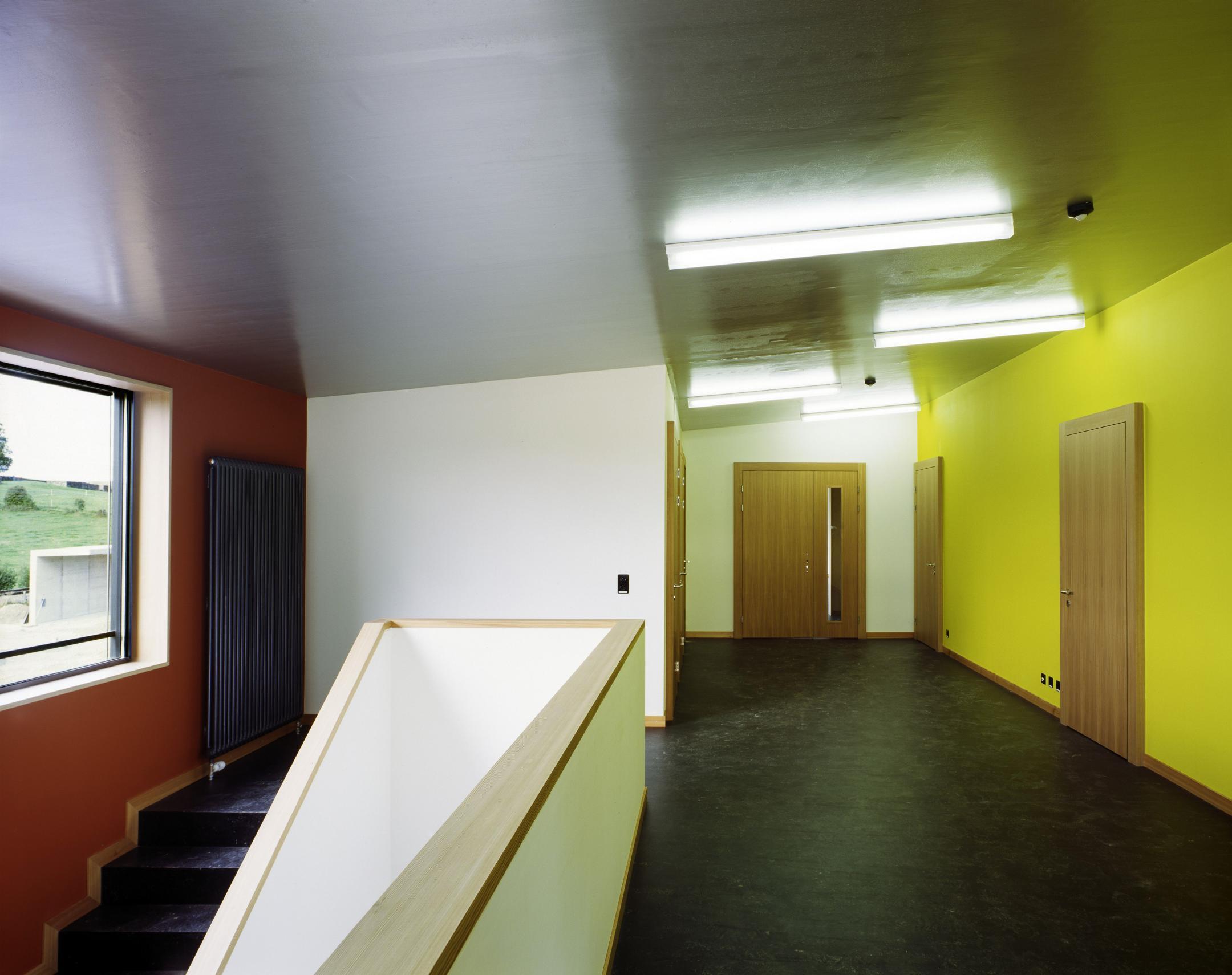 Korridor mit schwarzem Bodenbelag, gelben und roten Wänden und linksseitigem Treppenabgang.