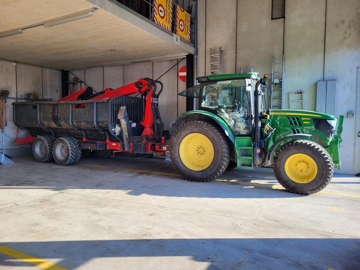 Abgestellter, roter Rungenwagen mit zusammengeklapptem Kran an einem grünen Traktor.