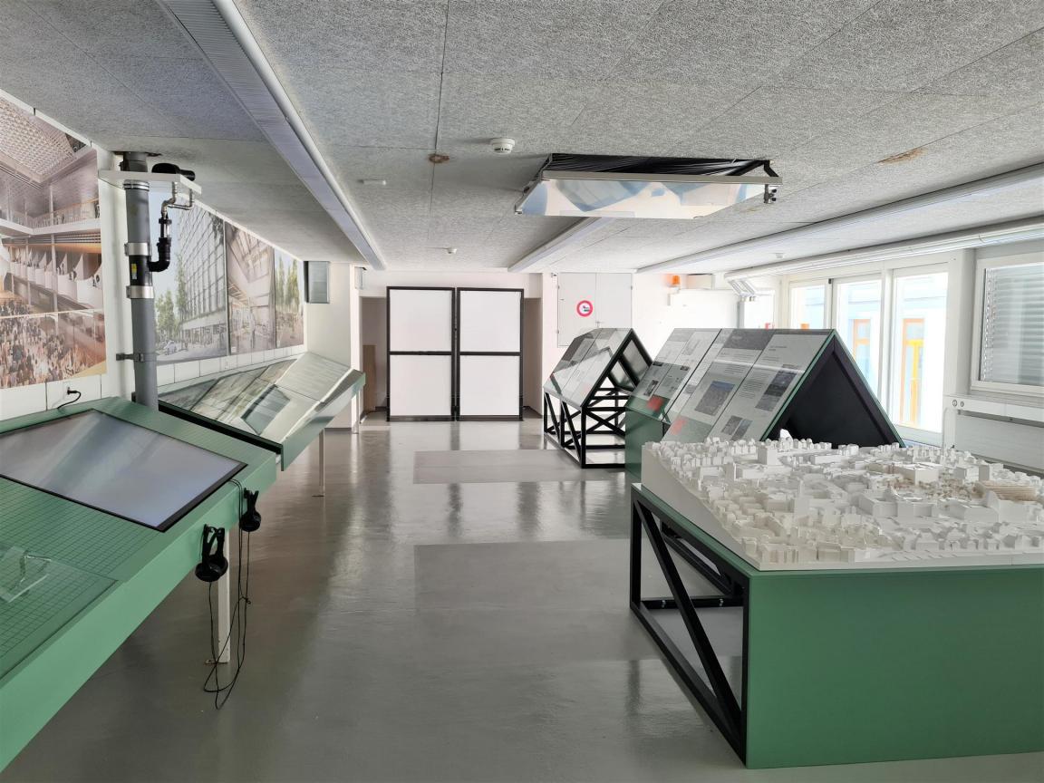 Schlichter Raum mit Ausstellung: Modell des Hochschulgebiets, Bildschirm mit Kopfhörern, Visualisierungen an den Wänden, grüne Konstruktionen mit Informationen