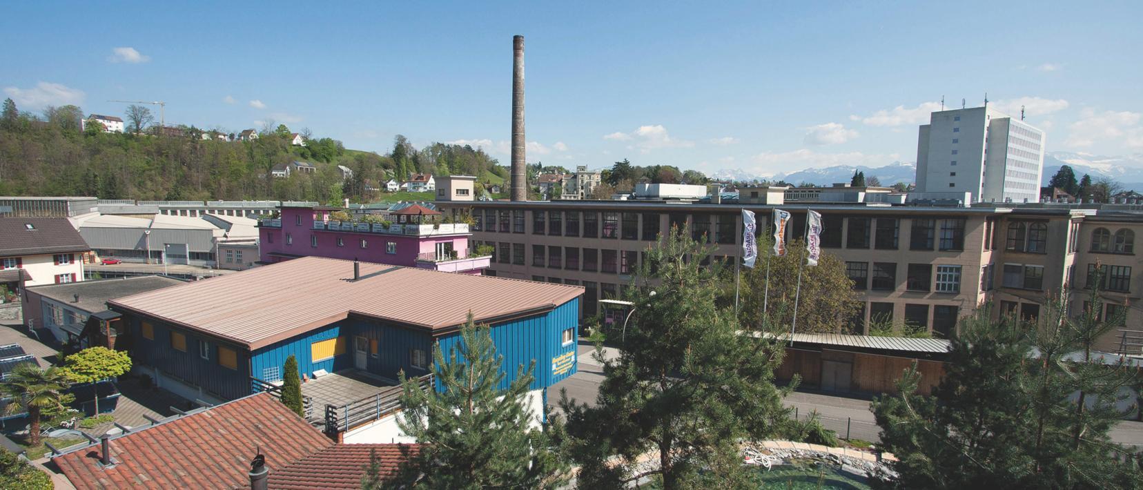 Blick auf das Joweid Technologie Zentrum Rüti. Im Vordergrund Bäume und ein blaues Nebengebäude, in der Bildmitte ein Hauptgebäude des ehemaligen Industrieareals.