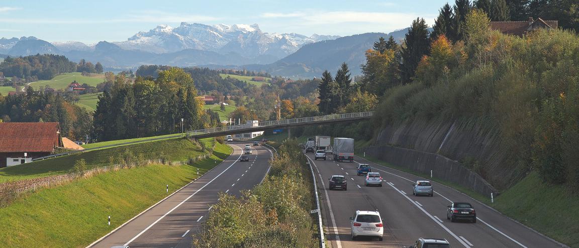 Blick gegen Osten von einer Brücke auf die beiden Fahrbahnen der Autobahn A3 zwischen Horgen und Wädenswil. Die Strasse führt durch eine sanfte Hügellandschaft mit Wiesen und Wäldern, am Horizont erhebt sich das Glärnischmassiv.