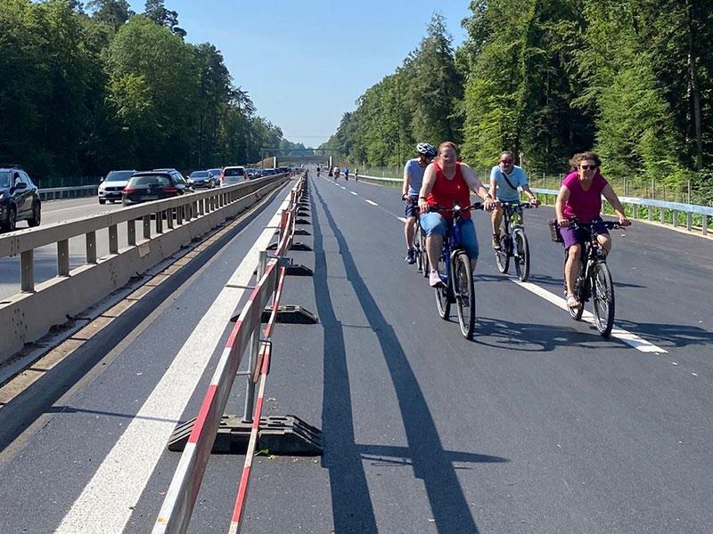 Eine breite, neu asphaltierte Strasse an einem sonnigen Tag. Im Vordergrund fahren mehrere Personen auf Fahrrädern der Kamera entgegen. Links trennt eine rot-wisse Absperrung die Fahrradspur von der Gegenfahrbahn, auf der sich Autos stauen. Die Strecke ist von dichten, grünen Wäldern gesäumt.
