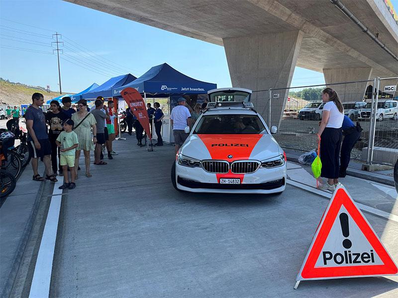 Ein weisses Polizeiauto der Marke BMW mit leuchtend orangeroter Front steht unter einer grossen Betonbrücke. Besucher, darunter Familien mit Kindern, stehen links daneben an Infoständen mit blauen Zelten. Im rechten Vordergrund warnt ein rot-weisses Faltsignal mit der Aufschrift «Polizei».