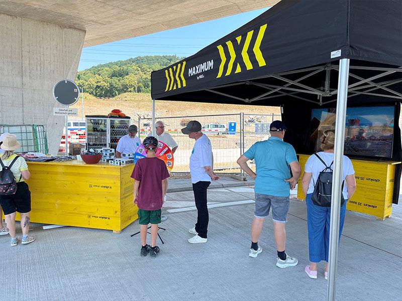Unter einer massiven Autobahnbrücke aus Beton befindet sich ein Ausstellungsstand mit der Aufschrift «MAXIMUM by HGC». Mehrere Personen stehen vor einer gelben Holztheke. Ein Junge mit Fahrradhelm schaut auf ein buntes Glücksrad. Rechts schauen Besucher auf einen grossen Bildschirm.