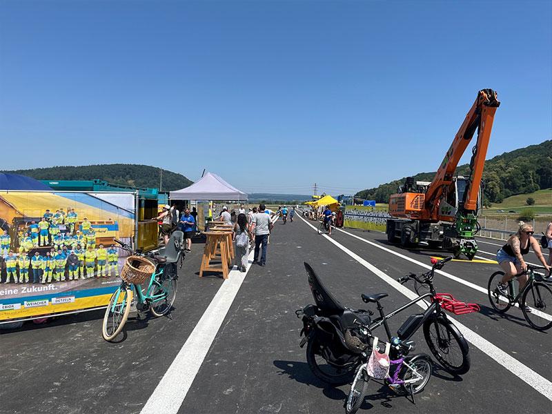 Eine weitläufige, frisch asphaltierte Strasse unter blauem Himmel. Auf der linken Seite stehen Infotafeln mit Teambildern, Stehtische und abgestellte Fahrräder. Auf der rechten Seite bewegt sich ein grosser, orangefarbener Kettenbagger. Im Vordergrund sind zwei Fahrräder mit Kindersitzen geparkt.
