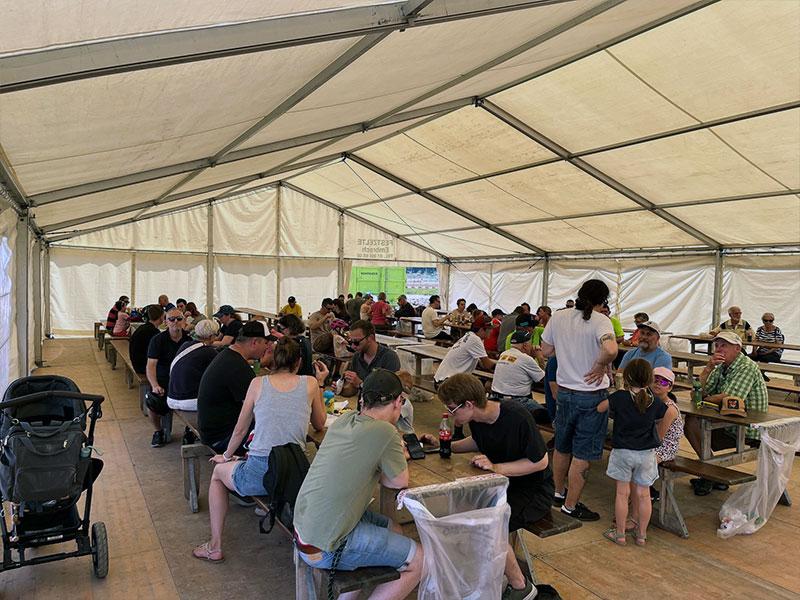 Das Innere eines grossen, hellen Festzelts mit einer Plane. Viele Menschen sitzen an langen Holztischen und Festbänken, essen, trinken und unterhalten sich. Im linken Vordergrund steht ein schwarzer Kinderwagen, im Zentrum blicken junge Männer auf ein Smartphone.