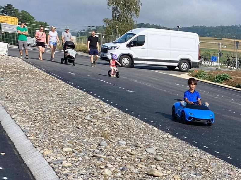 Eine asphaltierte Strasse im Aussenbereich. Im Vordergrund fährt ein kleiner Junge in einem blauen Spielzeug-Elektroauto, das einem Sportwagen nachempfunden ist. Dahinter folgt ein kleines Kind auf einem Laufrad mit violettem Helm. Im Hintergrund spaziert eine Gruppe von Erwachsenen neben einem weissen Lieferwagen.