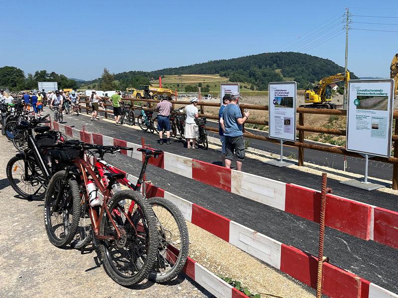 Ein abgesperrter Bereich neben einer Baustelle. Im Vordergrund stehen mehrere Mountainbikes. Dahinter betrachten Besucher weisse Informationstafeln, die an einem Holzgeländer befestigt sind. Auf einer Tafel steht gut lesbar «Umweltschonende Abwasserbehandlung». Im Hintergrund ist ein gelber Bagger zu sehen.