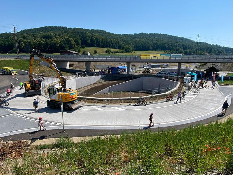 Blick von oben auf eine grosse, kreisrunde Betonanlage auf einer Baustelle mit einer tiefer gelegenen Zufahrt in der Mitte. Auf der oberen Ebene bewegen sich Radfahrer, Fussgänger und ein gelber Radlader. Im Hintergrund kreuzt eine Autobahnbrücke das Gelände, dahinter erstrecken sich grüne Hügel.