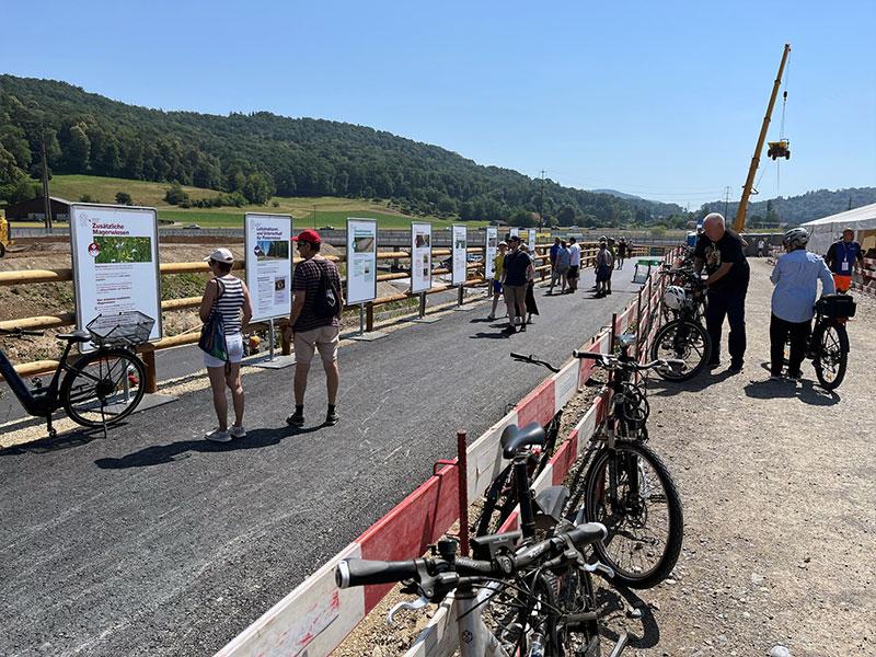 Eine Reihe von Informationstafeln an einem Holzgeländer entlang eines Weges. Die vorderste Tafel trägt den Titel «Zusätzliche Magerwiesen». Besucher stehen davor und lesen die Texte. Im Vordergrund ist der Lenker eines Fahrrads im Detail zu sehen, im Hintergrund ragt ein hoher gelber Baukran in den blauen Himmel.