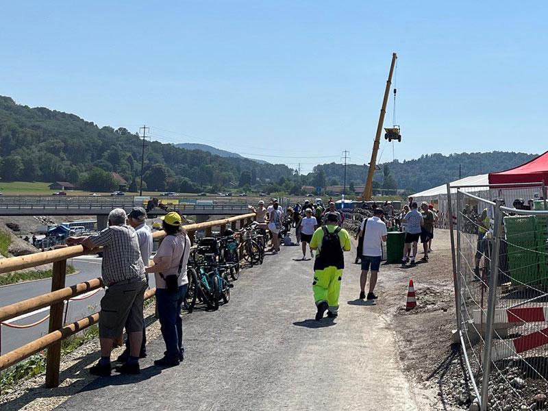 Ein Schotterweg am Rande einer Baustelle, auf dem sich viele Passanten und Bauarbeiter in Leuchtkleidung bewegen. Links stehen Menschen an einem Holzgeländer und schauen hinab. Im Hintergrund ist ein grosser, gelber Mobilkran zu sehen, der einen kleinen Muldenkipper hoch in der Luft an Seilen transportiert.