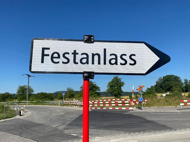 Ein grosses, weisses Pfeilschild mit schwarzem Rand und der Aufschrift «Festanlass» zeigt nach rechts. Es ist an einer leuchtend roten Stange befestigt. Im Hintergrund sieht man eine asphaltierte Strasse, rot-weiss gestreifte Absperrschranken, grüne Bäume und einen wolkenlosen, blauen Himmel.