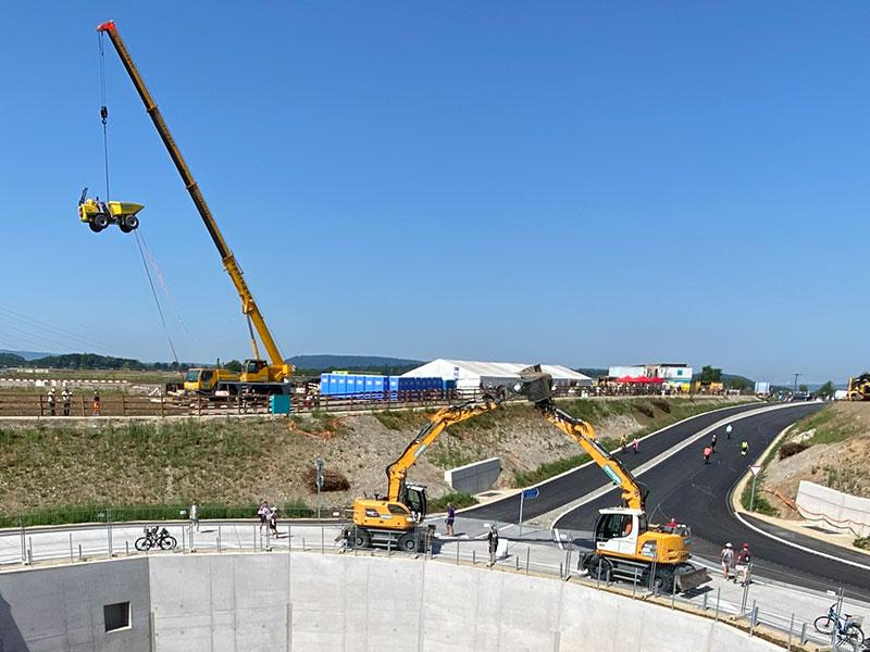 Eine Grossbaustelle unter strahlend blauem Himmel. Im Vordergrund stehen zwei gelbe Mobilbagger auf einer Betonfläche direkt an einer kreisrunden Vertiefung. Im linken Hintergrund ragt ein grosser, gelber Mobilkran empor, der einen kleinen Raddumper hoch in der Luft an Seilen hält. Einige Besucher beobachten die Szene.