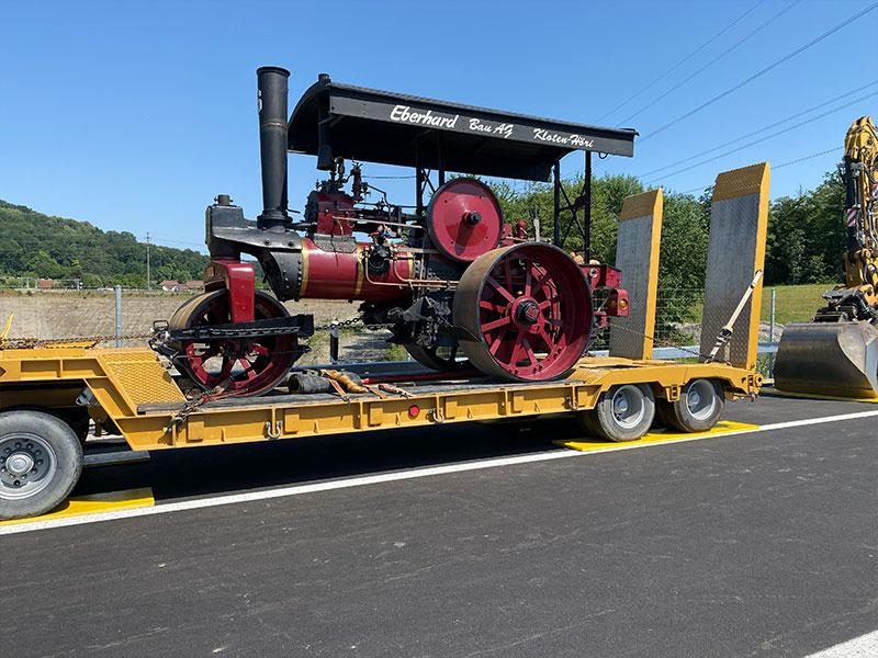 Eine historische, rot-schwarze Dampfwalze mit grossem Schornstein und einem Dach mit der Aufschrift «Eberhard Bau AG Kloten-Höri». Die Maschine ist mit Ketten auf einem gelben Tieflader-Anhänger gesichert, der auf einer frisch asphaltierten Strasse steht.