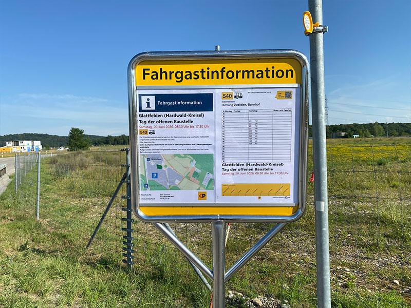 Eine Nahaufnahme eines gelb umrandeten Infokastens aus Metall, der an einem Pfosten montiert ist. Der Aushang trägt den Titel «Fahrgastinformation» und enthält Fahrpläne der Linie 540 sowie eine Umgebungskarte für den «Tag der offenen Baustelle» am Samstag, 20. Juni 2026. Im Hintergrund erstreckt sich eine Wiese mit gelben Blumen unter blauem Himmel.