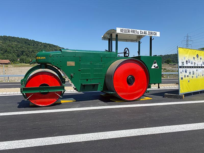 Eine klassische, dunkelgrüne Strassenwalze der Marke Ammann mit auffallend grossen, leuchtend roten Rädern. Auf dem weissen Dach steht der Firmenname «KELLER-FREI & Co. AG ZÜRICH». Die Walze steht auf neuem Asphalt neben einer gelben Informationstafel, im Hintergrund sind bewaldete Hügel erkennbar.