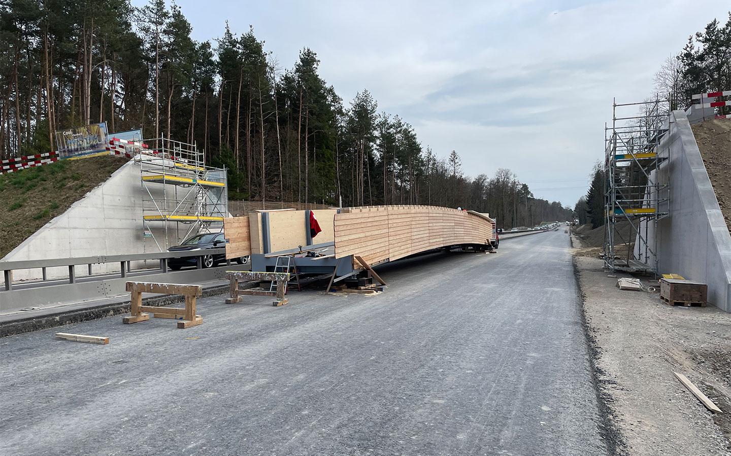 Ein hölzernes Brückenelement auf einem Spezialtransporter steht quer auf einer gesperrten, asphaltierten Strasse. Links und rechts der Fahrbahn befinden sich bereits betonierte Brückenwiderlager mit Gerüsten. Im Hintergrund ist ein dichter Wald unter einem bewölkten Himmel zu sehen.