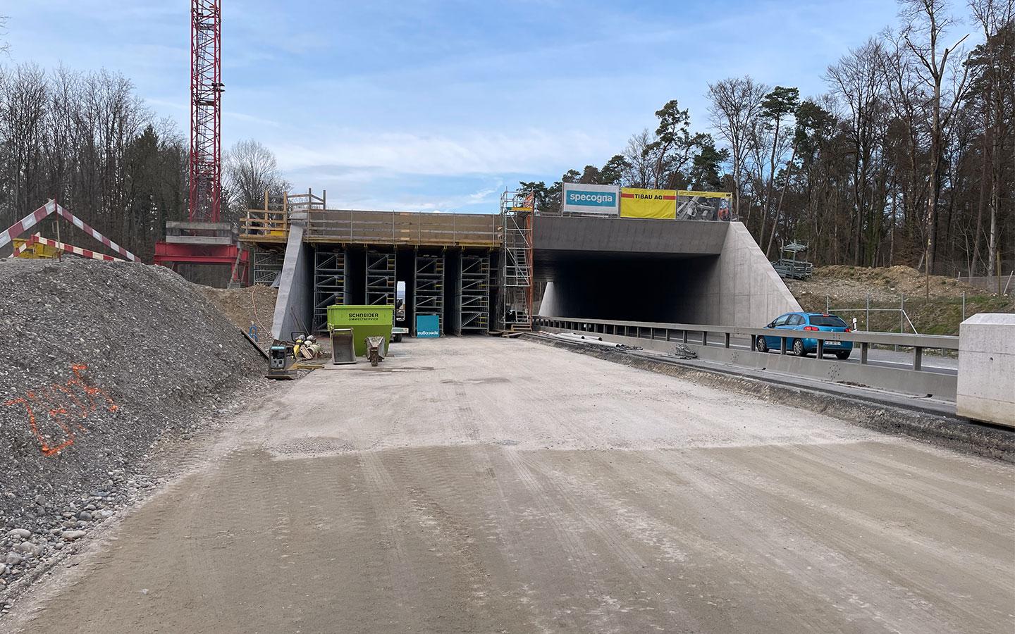 Baustelle einer Betonunterführung an einer Landstrasse. Die linke Röhre ist noch mit Gerüsten und Verschalungen ausgefüllt, während die rechte Röhre bereits für den Verkehr freigegeben scheint. Ein blauer PW fährt rechts vorbei. Im Hintergrund sind ein roter Baukran und ein Waldrand unter leicht bewölktem Himmel zu sehen.