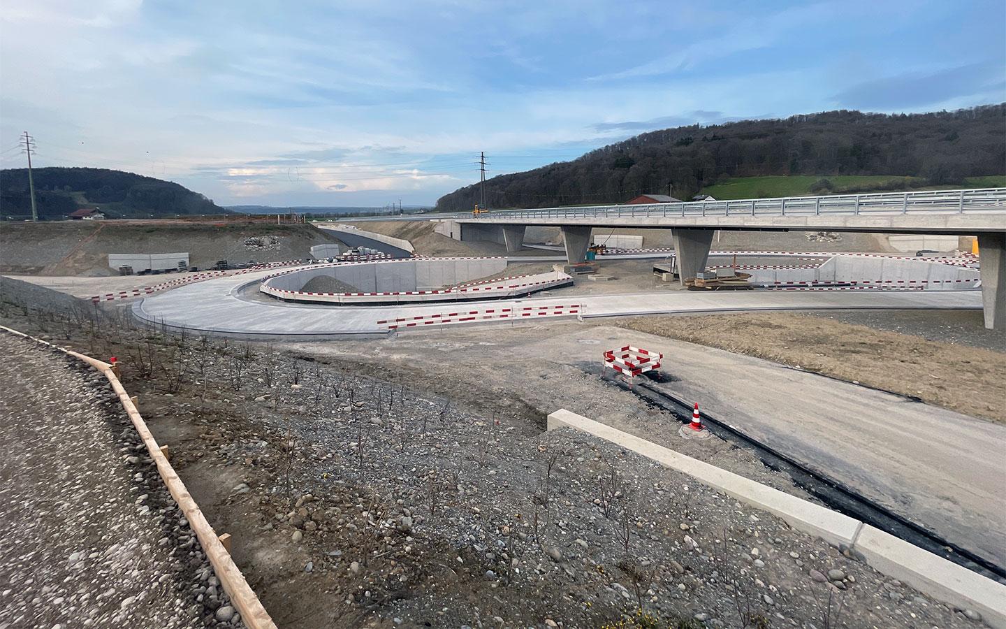 Weitwinkelaufnahme der grossen Strassenbaustelle im Hardwald. Im Vordergrund sind geschwungene Auffahrtsrampen aus Beton und frisch angelegte Böschungen zu sehen. Eine moderne Betonbrücke auf Pfeilern überspannt das Gelände. Im Hintergrund erstreckt sich eine hügelige, bewaldete Landschaft unter einem weiten, leicht bewölkten Himmel.