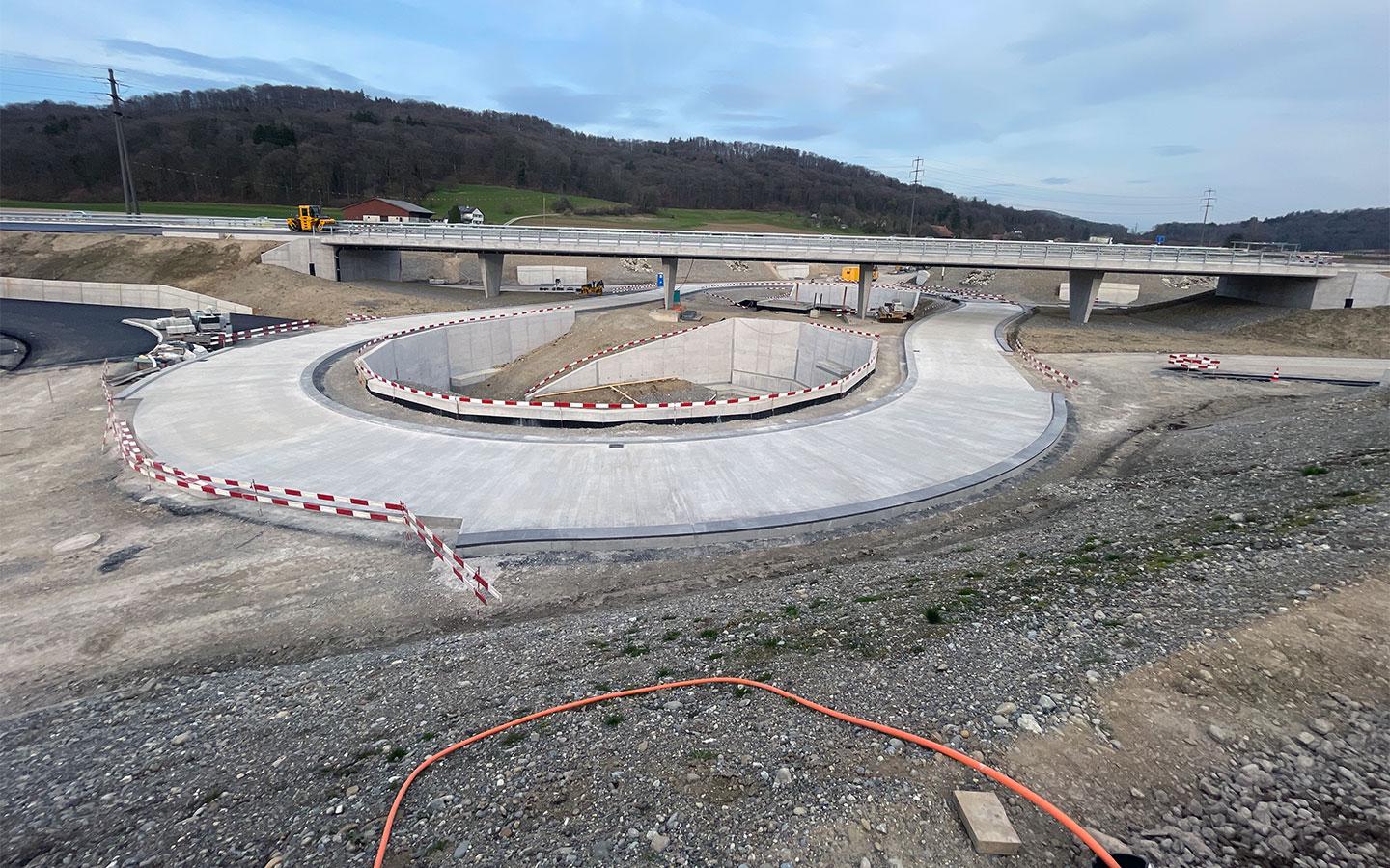 Totale einer grossflächigen Strassenbaustelle. Im Zentrum befindet sich eine hufeisenförmig angelegte Auffahrtsrampe aus hellem Beton, die von rot-weissen Absperrungen gesäumt ist. Dahinter verläuft eine lange Betonbrücke auf mehreren Pfeilern. Im Hintergrund sind bewaldete Hügel und eine weite Landschaft unter bewölktem Himmel zu sehen. Im Vordergrund liegt grober Kies mit einem orangefarbenen Kabel.