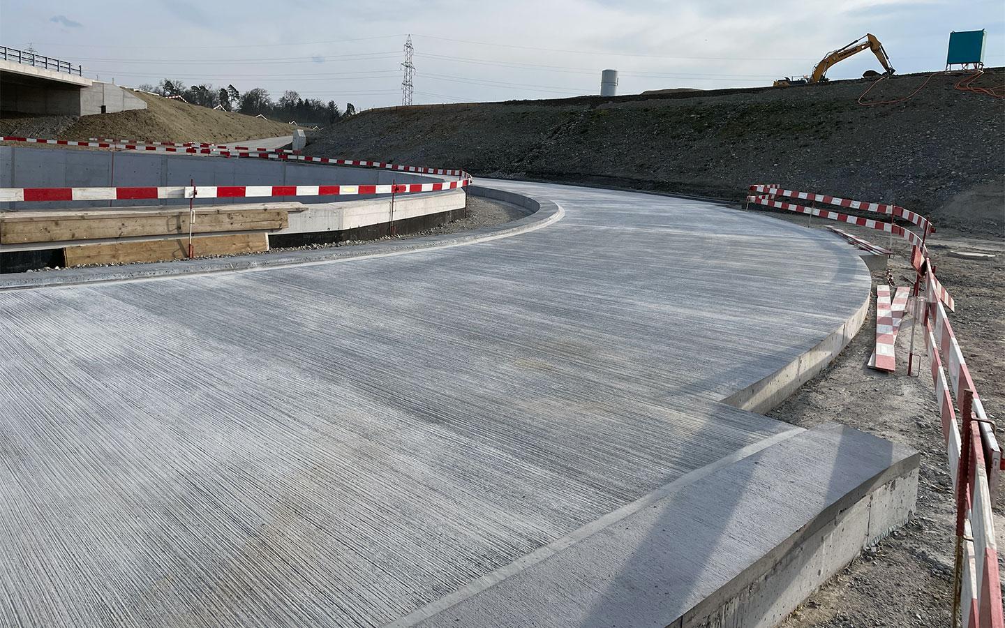 Nahaufnahme einer frisch betonierten, geschwungenen Fahrbahn. Die Betonoberfläche weist eine deutlich sichtbare, gerillte Textur auf. Die Fahrbahn wird von rot-weissen Baustellenabsperrungen und Betonelementen begrenzt. Im Hintergrund sind ein aufgeschütteter Erdhügel und ein gelber Bagger zu erkennen.