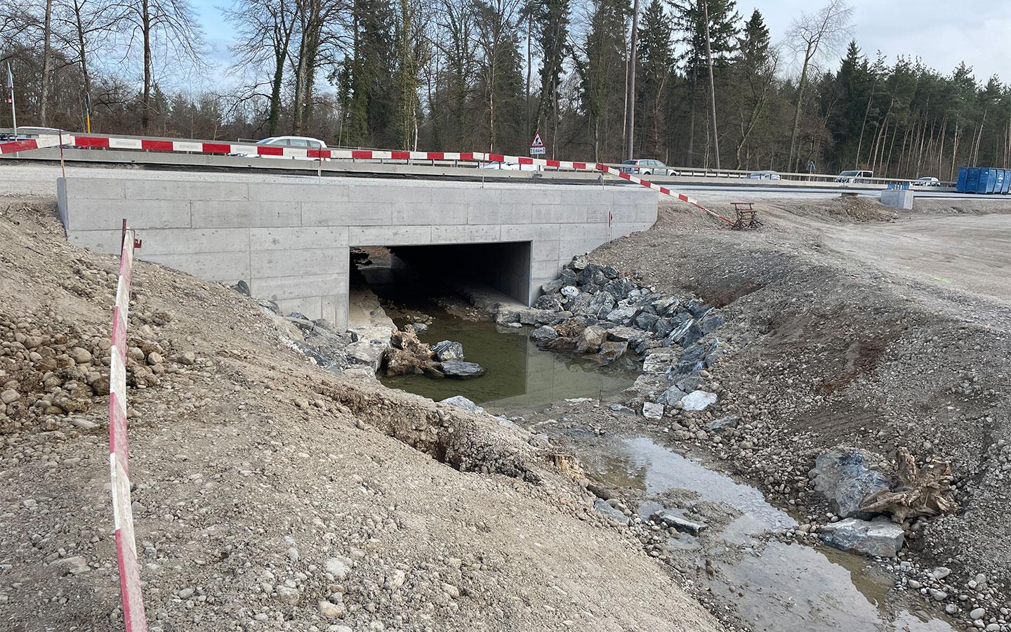 Ein massiver Beton-Durchlass unter einer aufgeschütteten Strasse. Durch die Unterführung fliesst ein kleiner Bach, dessen Ufer mit grossen Natursteinen befestigt ist. Im Hintergrund verläuft eine bereits für den Verkehr freigegebene Strasse mit Leitplanken, dahinter ist ein bewaldeter Hang zu sehen. Die gesamte Umgebung ist von Baustellen-Absperrband umgeben.