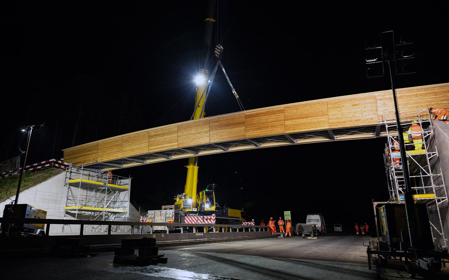 Ein grosser gelber Kran schwebt über einer Strasse und hält ein langes Brückenelement aus Holz und Stahl in der Luft, das zwischen zwei Betonrampen platziert wird.
