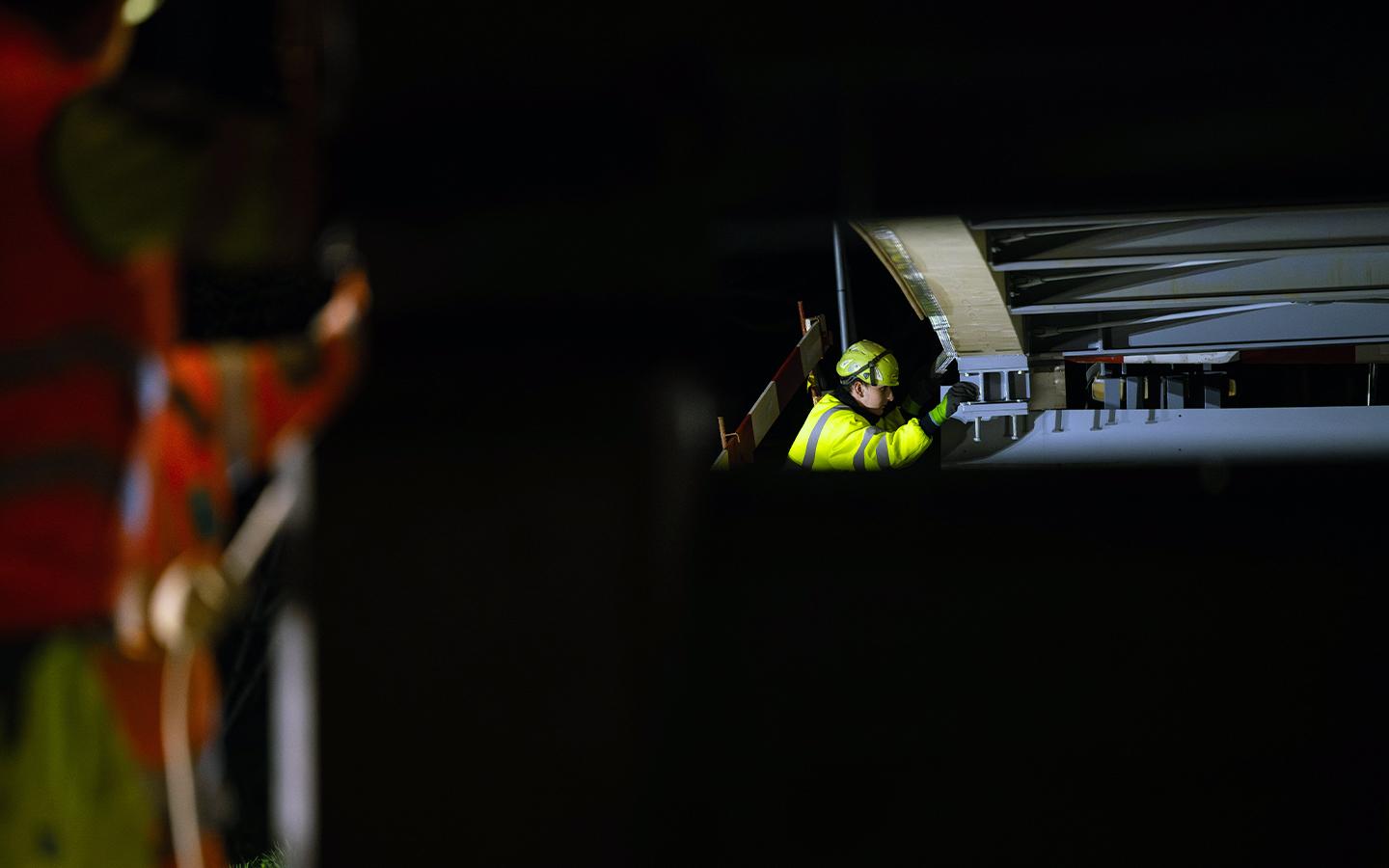 Nahaufnahme eines Bauarbeiters in neongelber Warnjacke und Helm, der nachts an der Unterseite einer Brückenkonstruktion arbeitet. 