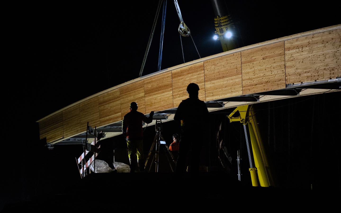 Blick auf die geschwungene Brücke. Die Seitenwände sind mit hellem Holz verkleidet. Im Hintergrund leuchten die Scheinwerfer des Krans.