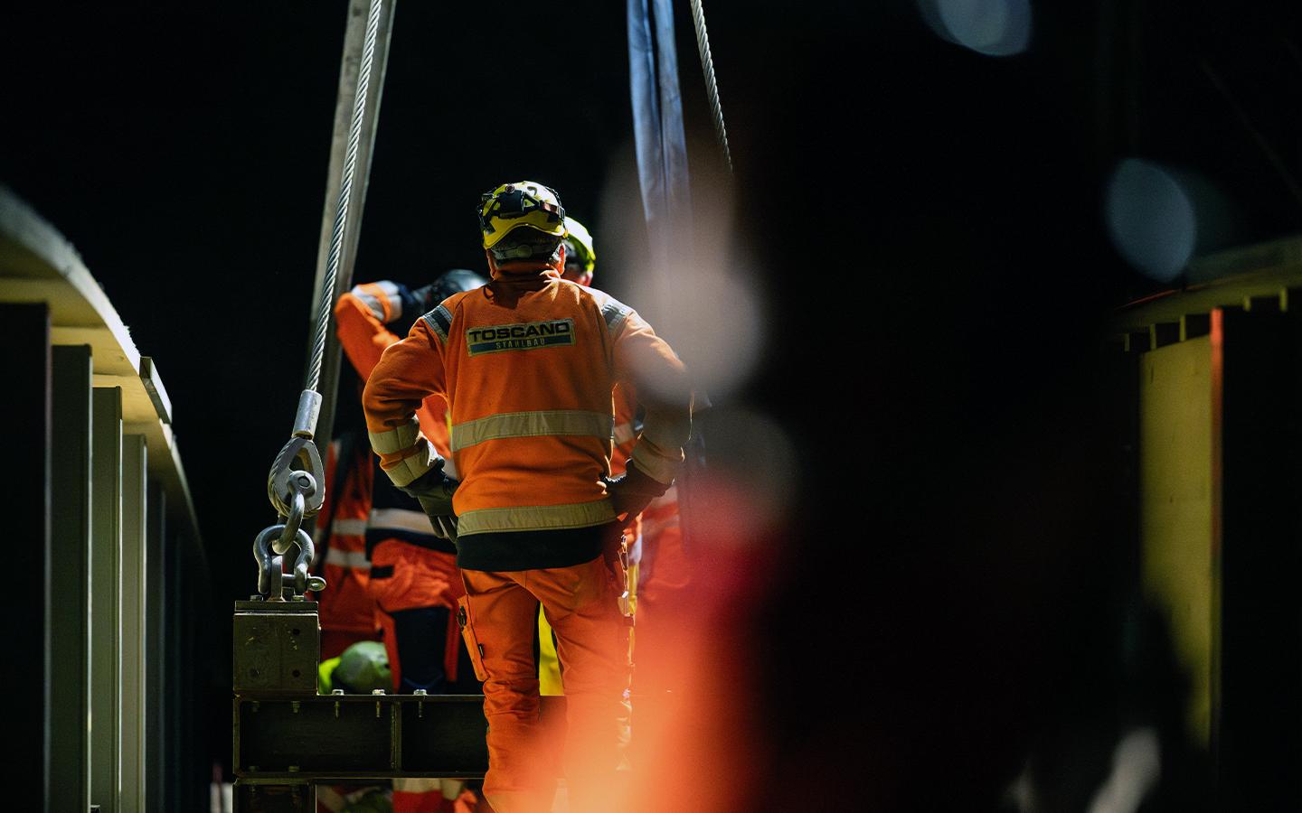 Ein Arbeiter in orangefarbener Montur mit dem Rücken zur Kamera beobachtet den Einhub. Im unscharfen Vordergrund und Hintergrund sind weitere Teammitglieder und die Stahlseile der Hebevorrichtung zu sehen.