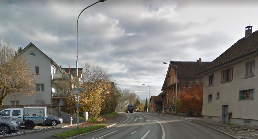Abschnitt der Dorfstrasse durch Obfelden, links und rechts stehen ein paar Wohnhäuser. Ein Lastwagen fährt über die Strasse. Am linken Strassenrand befindet sich ein Parkplatz mit Fahrzeugen.