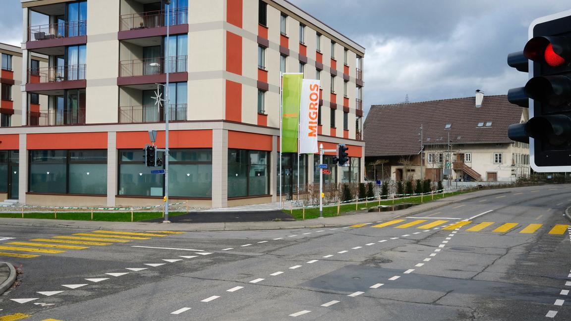 Kreuzung mit einer Supermarkt-Filiale an der Ecke eines modernen Gebäudes. Ein rotes Ampelsignal ist sichtbar. Im Hintergrund ist ein älteres Gebäude zu sehen. Die Strasse ist frisch markiert.